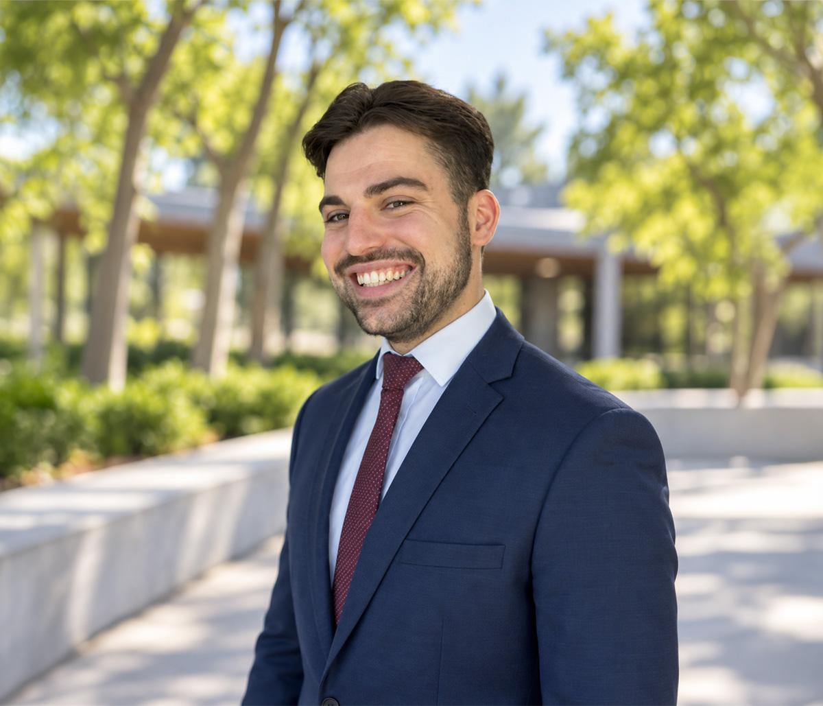 Joel Hasler, smiling, in a navy suit and dark red tie, in front of trees and a low glass building.