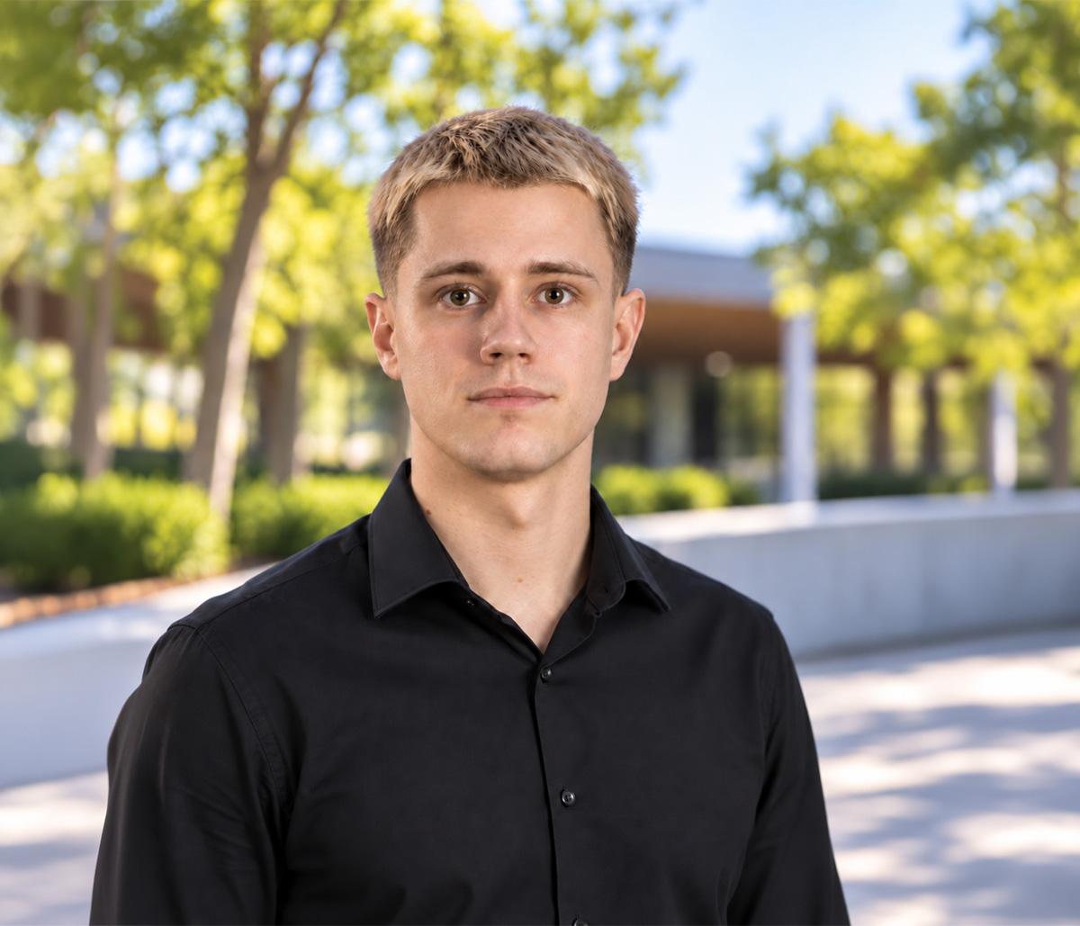Joey Merk, in a black shirt, in front of trees and a low glass building.